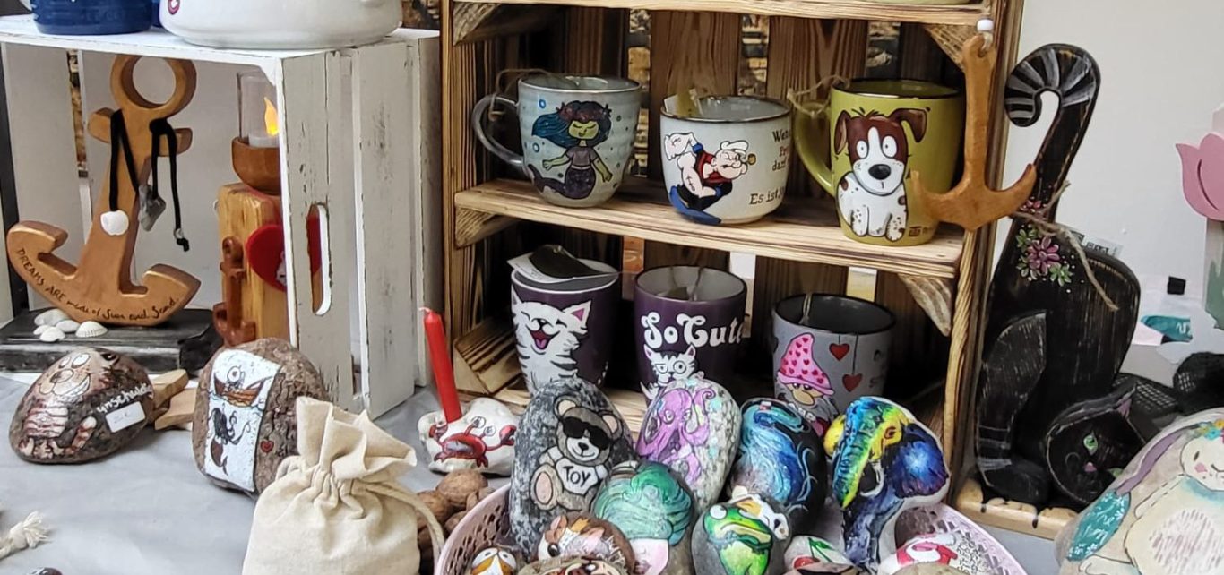 Ein Blick auf den Marktstand - Handgefertigte Holzskulpturen wie Leuchttürme, Kerzenständer und Anker neben handbemalten Tassen mit lustigen Motiven und Sprüchen und handbemalten Steine Handgefertigte Holzskulpturen wie Leuchttürme, Kerzenständer und Anker sind neben handbemalten Tassen mit lustigen Motiven und Sprüchen und handbemalten Steinen zu sehen